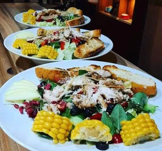 سالاد سزار مرغ تازه کافه رستوران مکث: سه پرس جذاب با مرغ گریل، ذرت، سیب و نان تست بر روی میز چوبی.