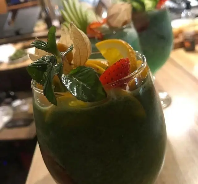 نوشیدنی اسموتی سبز و طبیعی کافه رستوران مکث با گارنیش میوه تازه شامل نعنا، فیسالیس، پرتقال و توت فرنگی.