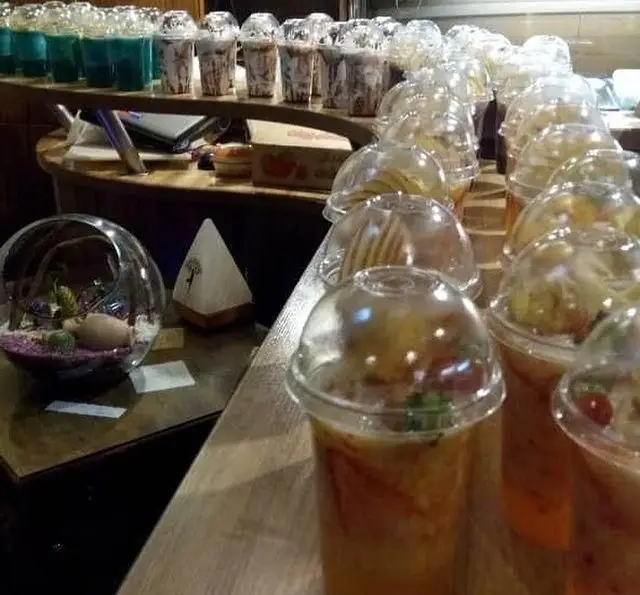 کافه رستوران مکث: میز پذیرایی پر از انواع نوشیدنی‌های خنک، اسموتی‌های میوه‌ای رنگارنگ، قهوه و دسرهای لذیذ در ظروف یکبار مصرف، با تزئین دکوراتیو.