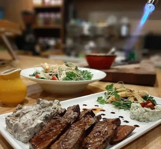 استیک لذیذ با سس مخصوص و سالاد تازه، در کافه رستوران مکث (Maks) سرو شده است. یک سرآشپز در حال تکمیل آماده‌سازی غذا با مشعل است.