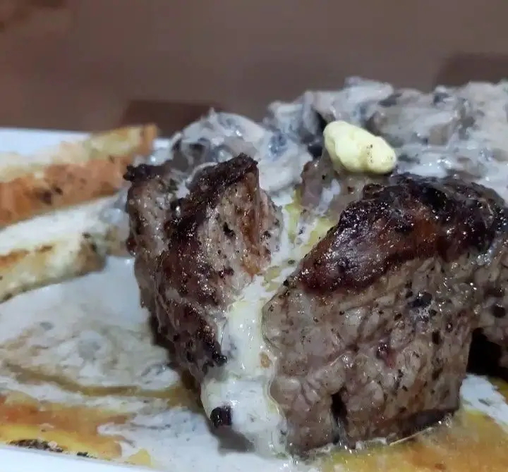 استیک آبدار کافه رستوران مکث با سس قارچ خامه‌ای و تکه‌ای کره ذوب شده، در کنار نان تست، یک غذای لذیذ برای تجربه در مکث.