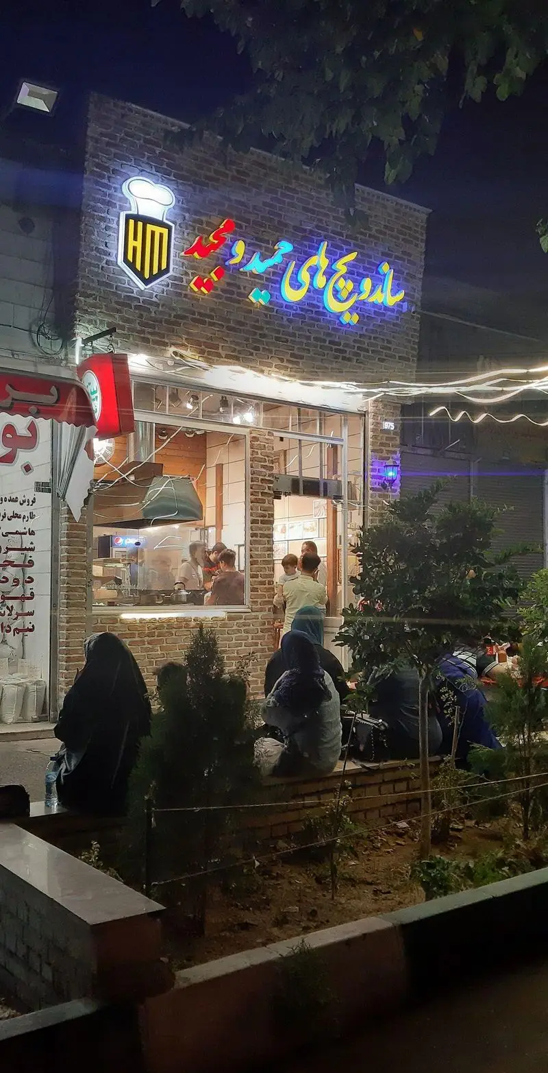 نمای جذاب شبانه فست فود و ساندویچی حمید و مجید با تابلوی نئون درخشان، مشتریان و پرسنل فعال در آشپزخانه آجری.