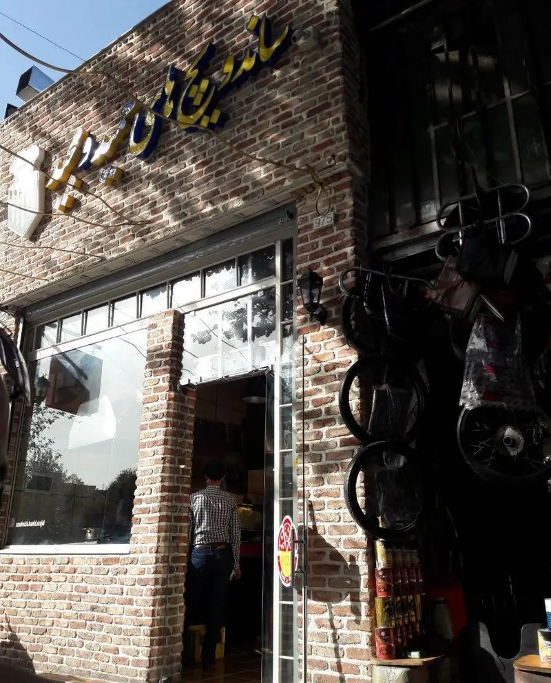 نمای بیرونی جذاب فست فود حمید و مجید با تابلوی برجسته زرد و آبی روی دیوار آجری سنتی. ورودی فعال رستوران.