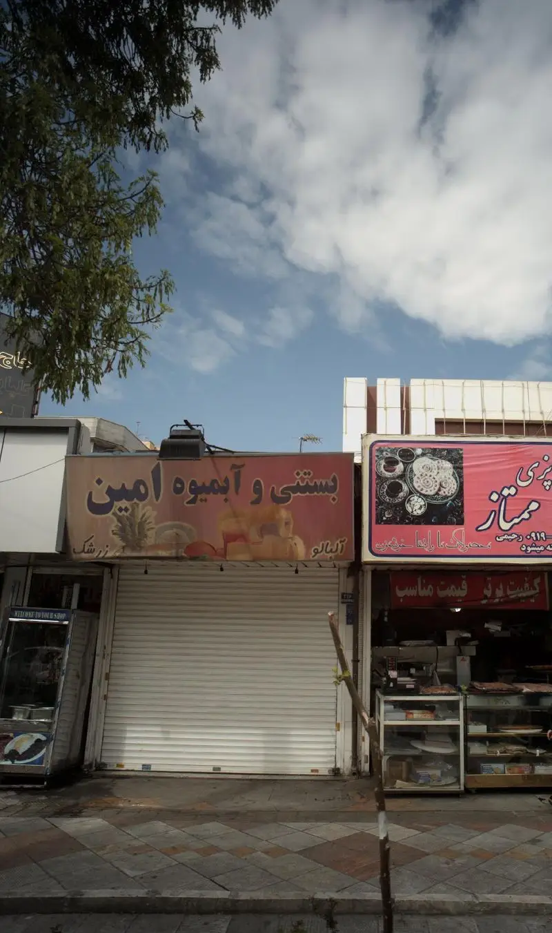 مغازه بستنی و آبمیوه امین با تابلوی قرمز رنگ و کرکره بسته، کنار شیرینی فروشی ممتاز. نمایی از خیابان و آسمان آبی با ابرها.