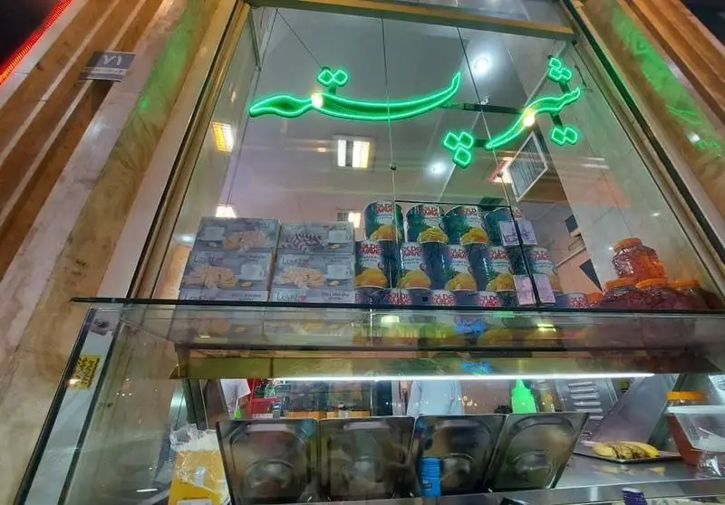 تصویری از نمای شب بستنی مسعود، شامل ویترین پر از محصولات متنوع و تابلوی نئونی سبز "بستنی مسعود" در بالای آن.