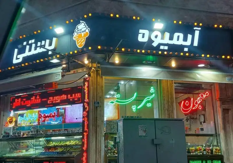 نمایی از مغازه بستنی مسعود با تابلوهای روشن بستنی، آبمیوه، شیرپسته و معجون. ویترین پر از میوه و دسرهای خنک.