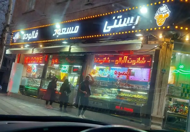 نمای شبانه و پرنور بستنی و فالوده مسعود، با تابلوهای روشن بستنی، فالوده، آبمیوه و حضور مشتریان در حال خرید.