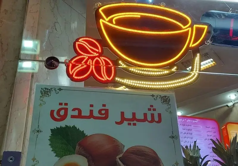 تابلو نئون درخشان بستنی مسعود با طرح فنجان قهوه و دانه های قهوه درخشان، در کنار بنر معرفی شیر فندق. تصویری جذاب از ورودی کافه و تنوع نوشیدنی ها.