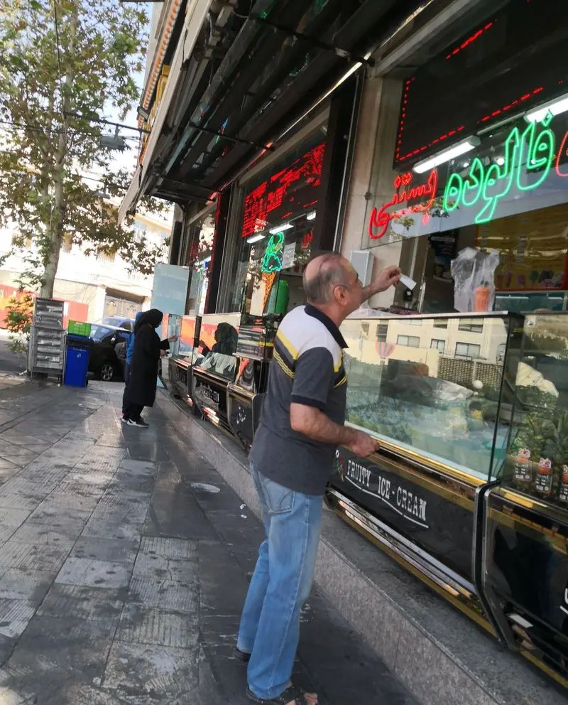 مردی در حال خرید از مغازه بستنی مسعود، با تابلوهای نئونی فالوده و بستنی روشن در ویترین بزرگ 'FRUITY ICE-CREAM' و مشتریان دیگر در پیاده‌رو.