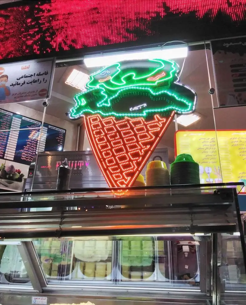 ویترین پر از انواع بستنی و دسرهای خوشمزه در بستنی مسعود، با تابلو نئون بزرگ بستنی قیفی نورانی و جذاب.