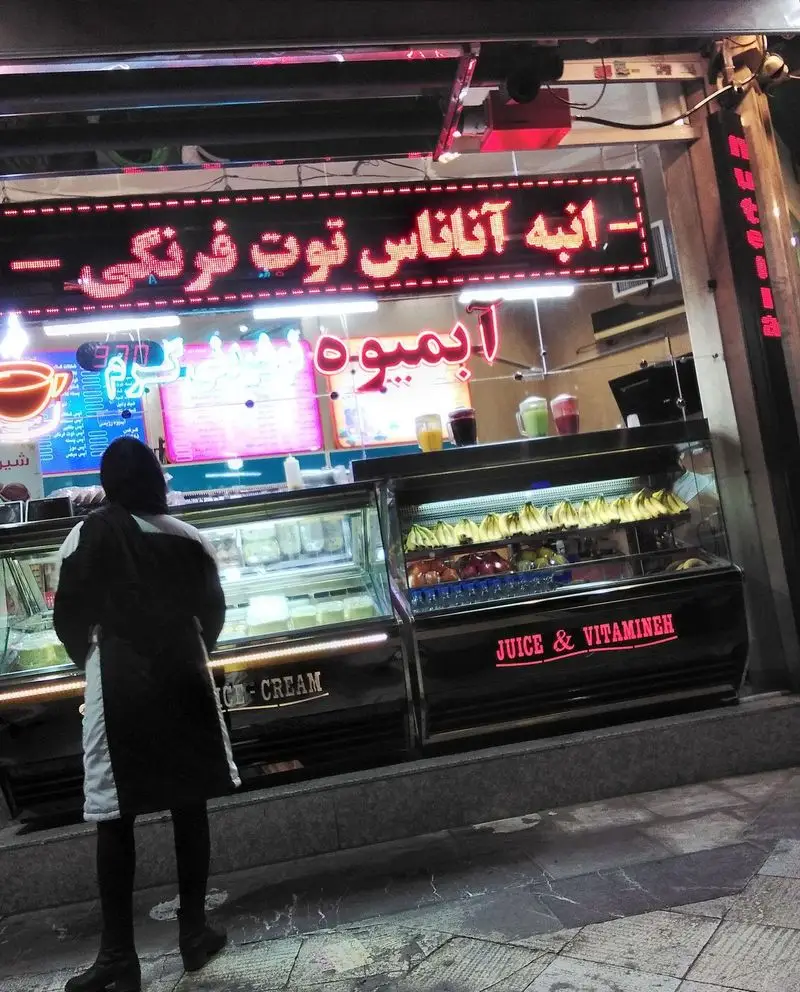 فروشگاه آبمیوه و بستنی مسعود در شب: ویترین پر از میوه، نوشیدنی‌های تازه (انبه، آناناس، توت فرنگی) و بستنی ویتامینه.