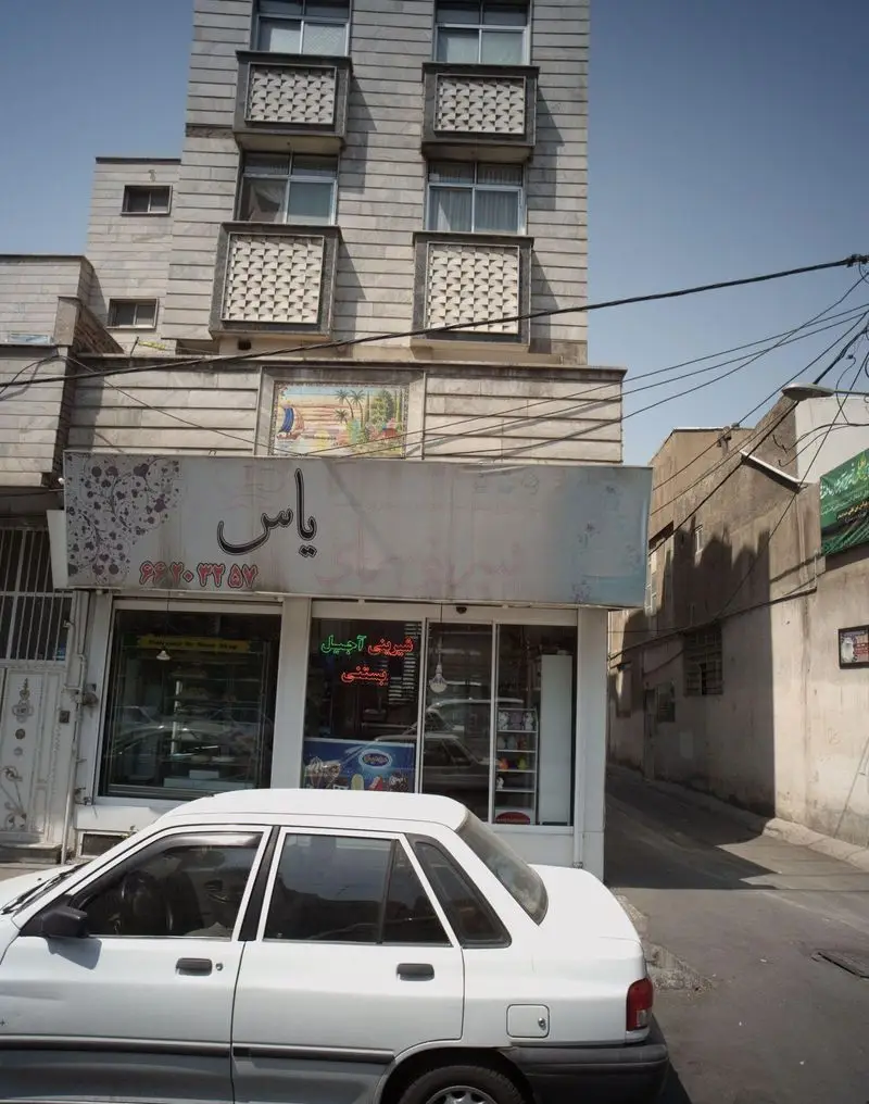 تصویر نمای بیرونی شیرینی سرای پدر، مکانی ایده‌آل برای خرید شیرینی، آجیل و بستنی، با خودروی سفید پارک شده و ساختمان چند طبقه.