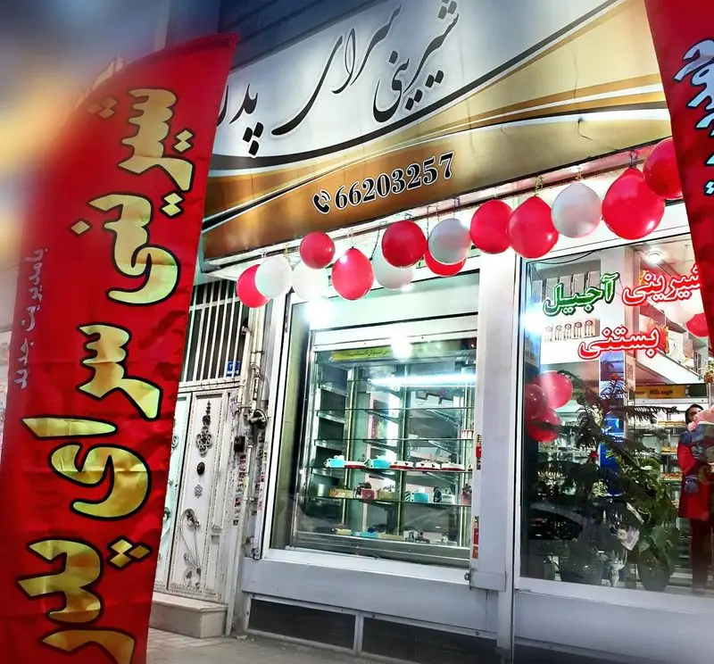 نمای بیرونی شیرینی سرای پدر با دکوراسیون جشن، بادکنک‌های قرمز و سفید، ویترین‌های پر از شیرینی، آجیل و بستنی.