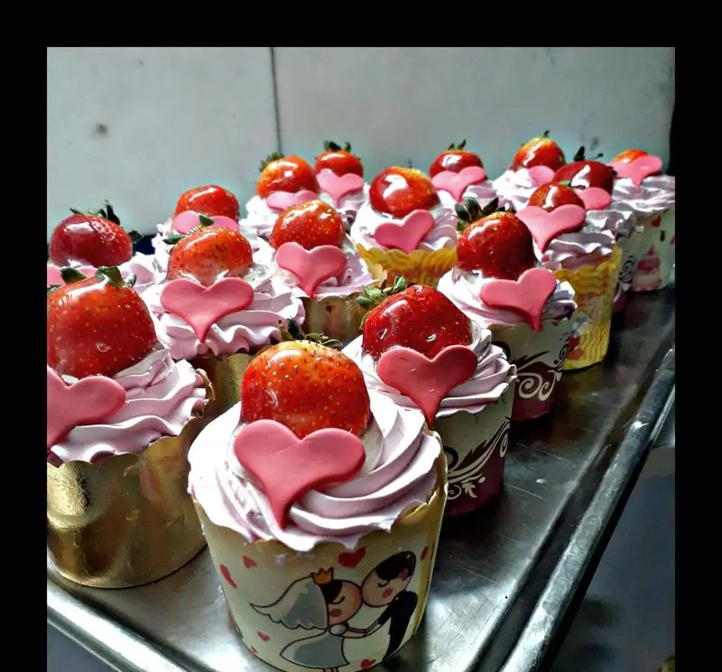 تصویر ردیفی از کاپ کیک‌های توت فرنگی تازه و خوشرنگ با تزیین خامه صورتی و قلب‌های زیبا، مناسب ولنتاین و جشن‌های عروسی از شیرینی سرای پدر.