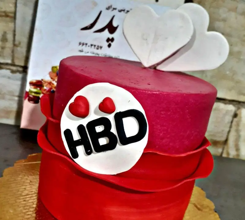 کیک تولد دو طبقه قرمز رنگ با تزیین قلب‌های سفید و نشان HBD، یک گزینه عالی از شیرینی سرای پدر برای جشن‌های شما.