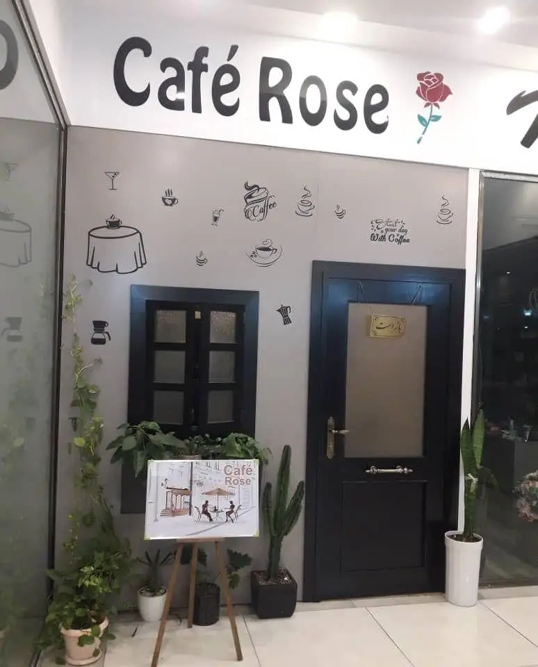ورودی دنج و زیبای کافه رز با دکوراسیون جذاب قهوه و گل رز، شامل پنجره مشکی، درب شیک، گیاهان و تابلوی هنری، فضایی دلنشین برای مشتریان.