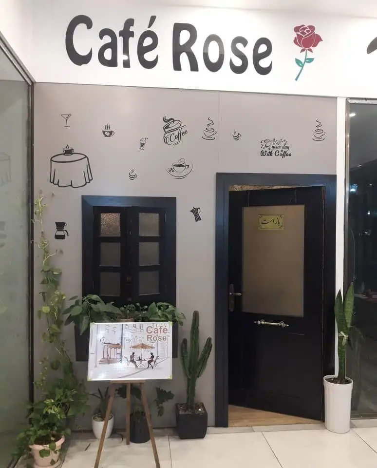 نمای بیرونی کافه رز با تابلو "Café Rose" و آرم گل رز، دیوارهای تزئین شده با نمادهای قهوه، در ورودی، پنجره و گیاهان سرسبز.