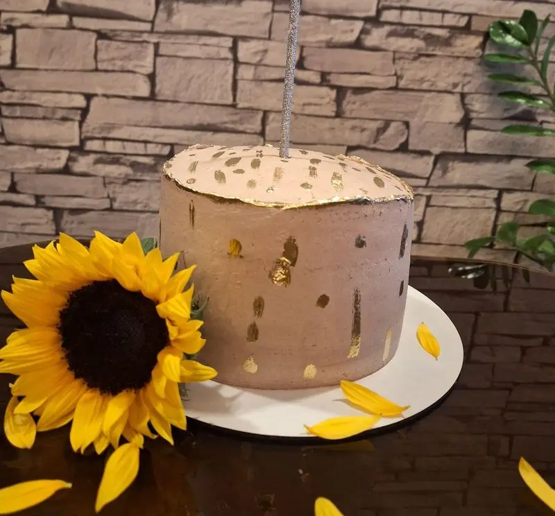 کیک تولد کافه رز با تزیین ورق طلا و شمع نقره‌ای. گل آفتابگردان و گلبرگ‌های زرد، میز شیشه‌ای و دیوار آجری. انتخابی خاص برای جشن‌ها.