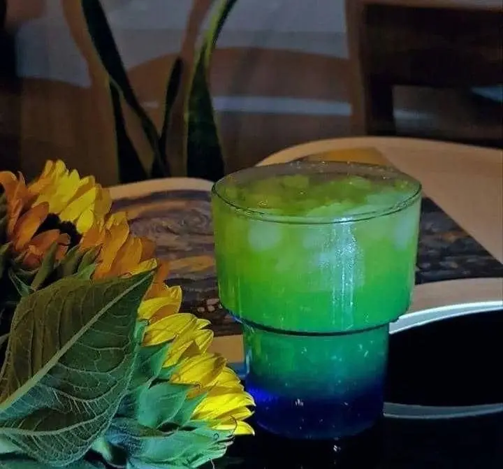 یک نوشیدنی سبز خنک کافه رز با یخ در لیوانی زیبا، کنار دسته گل های آفتابگردان و کتاب. فضایی دلنشین و تابستانی.