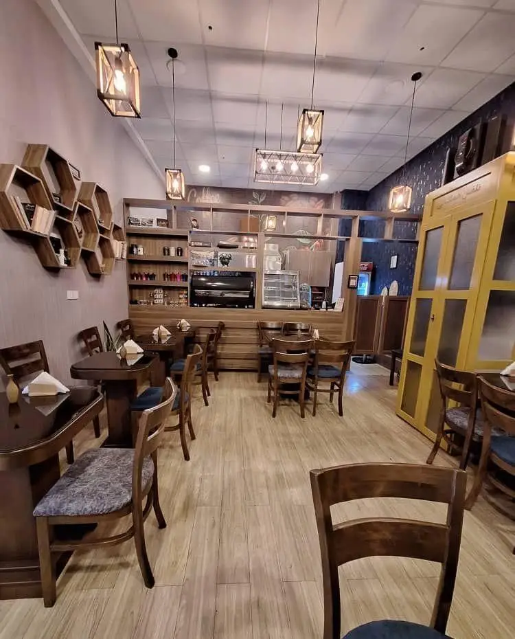 فضای داخلی دنج و شیک کافه رز با میز و صندلی‌های چوبی، قفسه‌های شش ضلعی و دکوراسیون گرم برای تجربه نوشیدنی‌های خوشمزه.