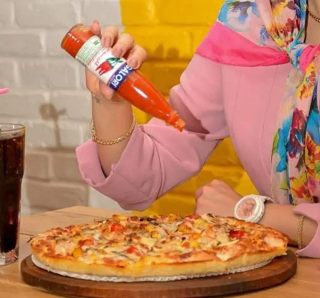 یک مشتری خوشحال در رستوران جیکی بیف در حال افزودن سس تند سالوری به پیتزای داغ و خوشمزه. تجربه‌ای لذیذ از غذای تازه.