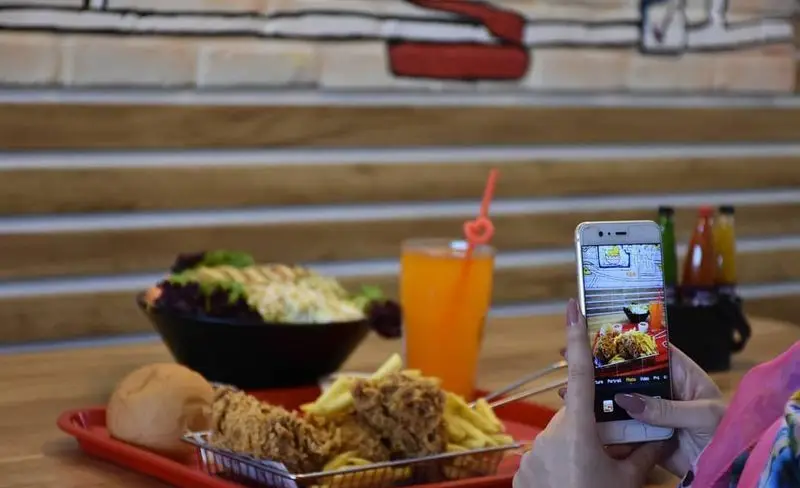 مشتری در رستوران جیکی بیف در حال عکاسی از غذای خوشمزه شامل مرغ سوخاری، سیب زمینی سرخ کرده، سالاد و نوشیدنی با موبایل است.