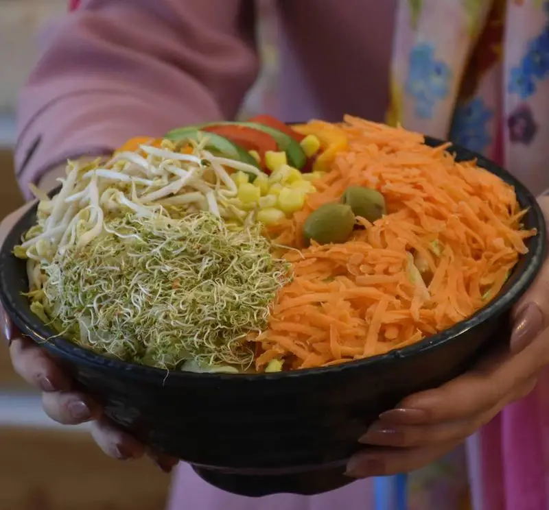 یک کاسه بزرگ سالاد تازه و رنگارنگ جیکی بیف شامل جوانه، هویج رنده شده، ذرت، خیار و زیتون برای یک وعده سالم و کامل.