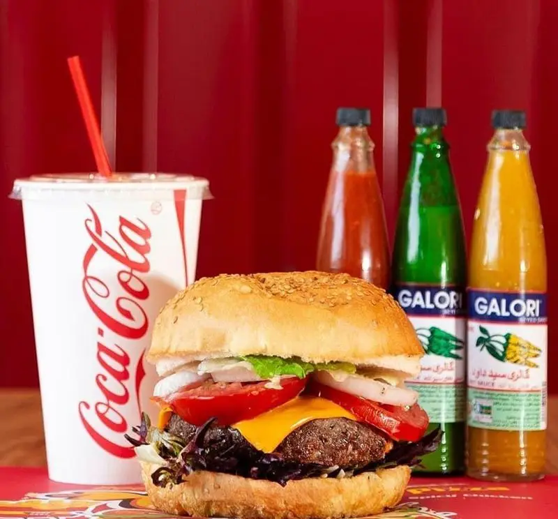 همبرگر بزرگ و آبدار جیکی بیف با پنیر ذوب شده، گوجه، پیاز و سبزیجات تازه، کنار نوشابه کوکاکولا و سس‌های متنوع گالوری.