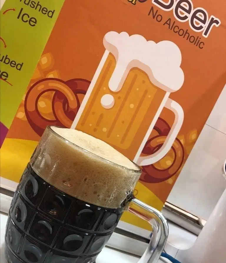 لیوان نوشیدنی ماءالشعیر جیکی بیف با فوم لذیذ و خنک، در مقابل پوستر آبجو بدون الکل با طرح چوب شور و گزینه‌های یخ.