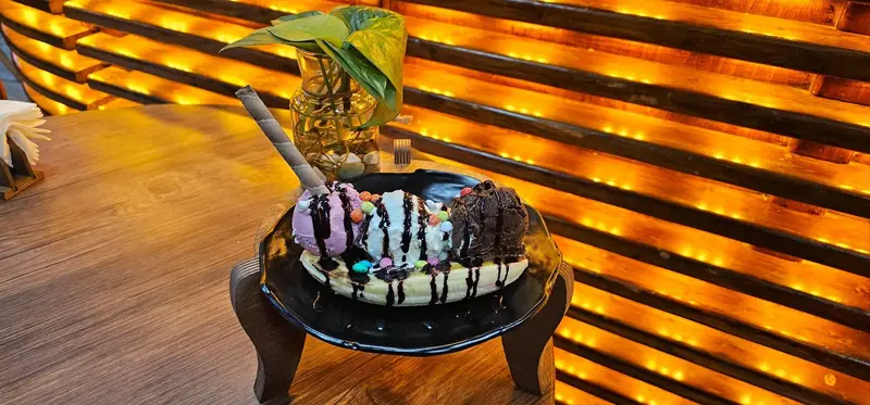 دسر محبوب بستنی موز کافه لوف شامل سه اسکوپ بستنی میوه‌ای و شکلاتی با سیروپ و آبنبات رنگی، مناسب برای لذت بردن در فضای دلنشین.