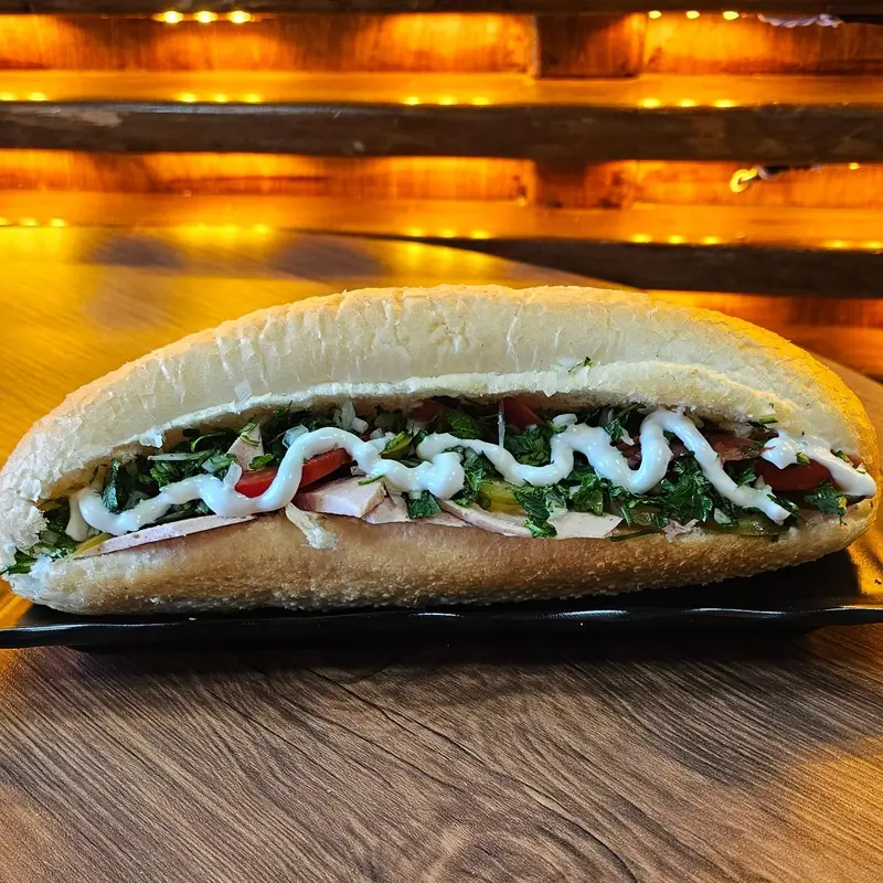 ساندویچ باگت تازه و پرملات کافه لوف با ژامبون، سبزیجات تازه، گوجه، خیارشور و سس مخصوص روی میز چوبی در محیط گرم کافه.