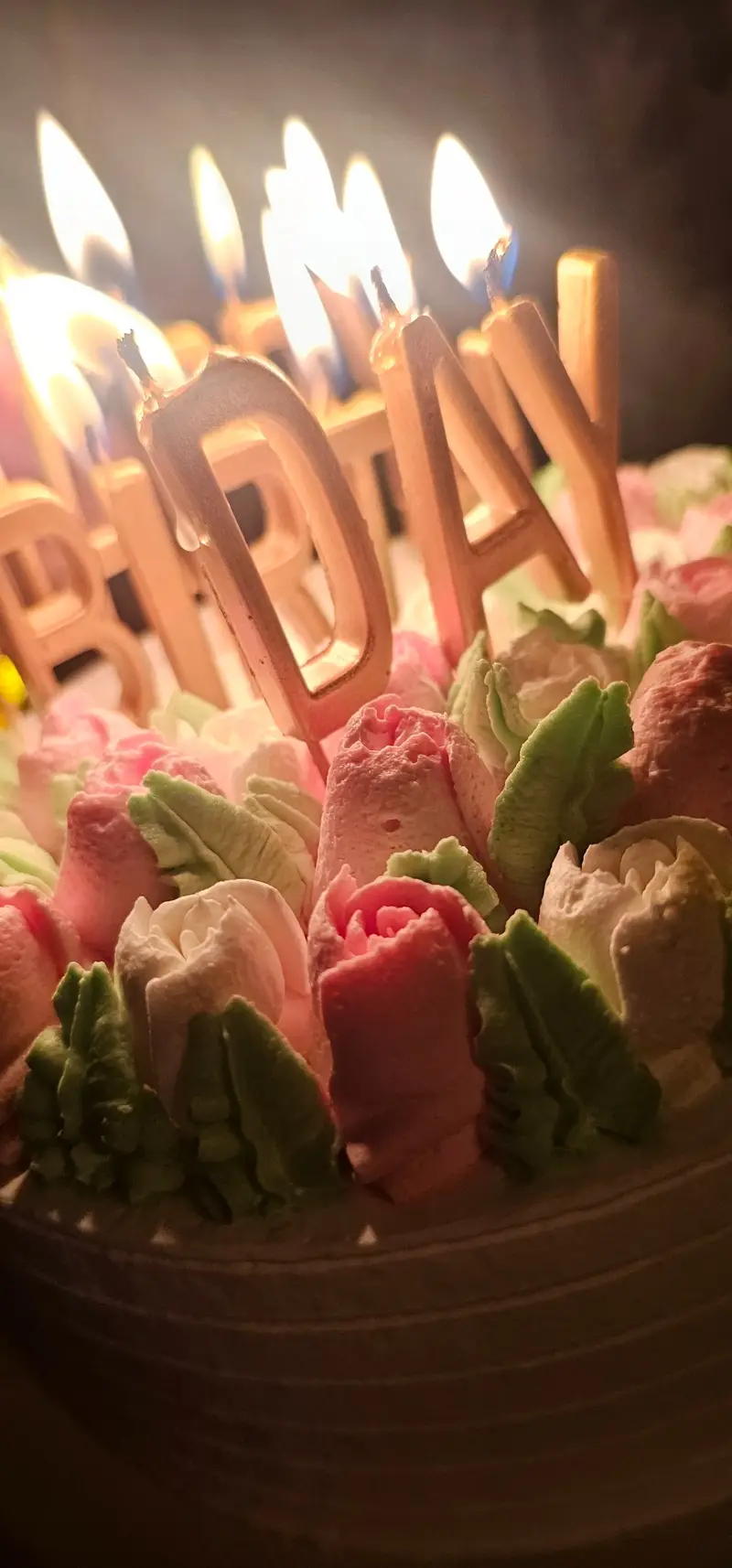 کیک تولد باشکوه کافه لوف با شمع‌های طلایی "BIRTHDAY" و شعله‌های درخشان، همراه با تزیینات گل‌های خامه ای صورتی و سبز. ایده‌آل برای جشن‌های تولد.