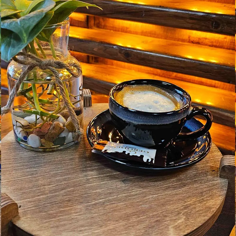 فنجان قهوه اسپرسو تازه و گرم با کرمای غلیظ در کافه لوف، همراه با شکلات و گلدان تزیینی روی میز چوبی، فضایی دلنشین و آرام برای لذت بردن از نوشیدنی.