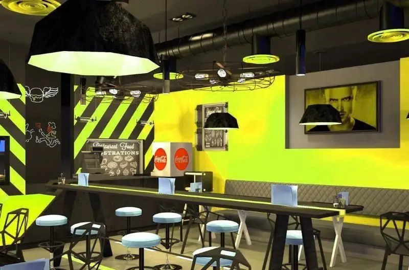 فضای داخلی مدرن و جذاب رستوران RAMCY با دکوراسیون صنعتی، دیوار زرد و مشکی، میزهای بلند و صندلی‌های شیک.