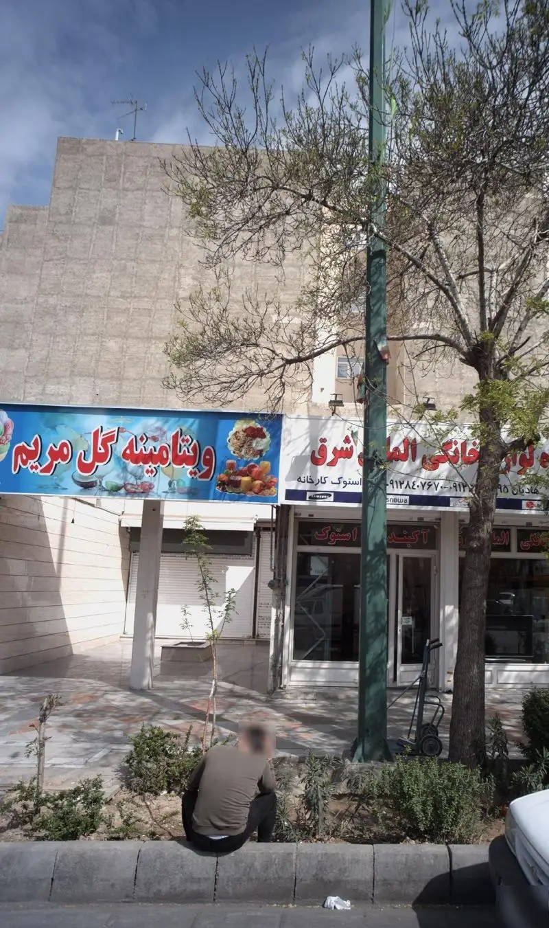 نمای خیابانی از مغازه ویتامینه گل مریم با تابلوی بزرگ آبی رنگ و درختان، نشان‌دهنده محیط شهری و کسب‌وکار پر جنب‌وجوش.