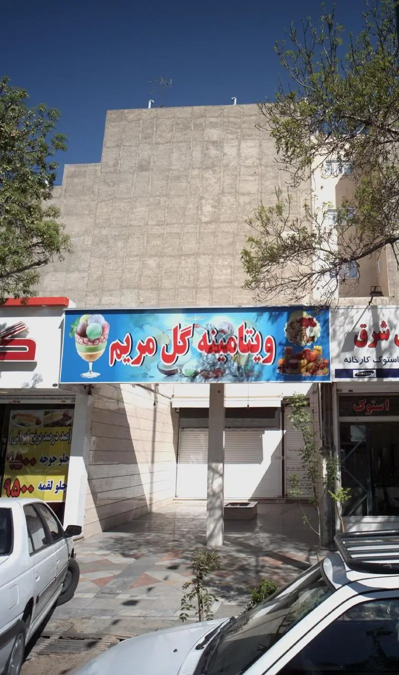 نمای بیرونی مغازه ویتامینه گل مریم با بنر آبی رنگ و تصویر بستنی و دسر، به همراه درختان و نمای خیابان در روز روشن.