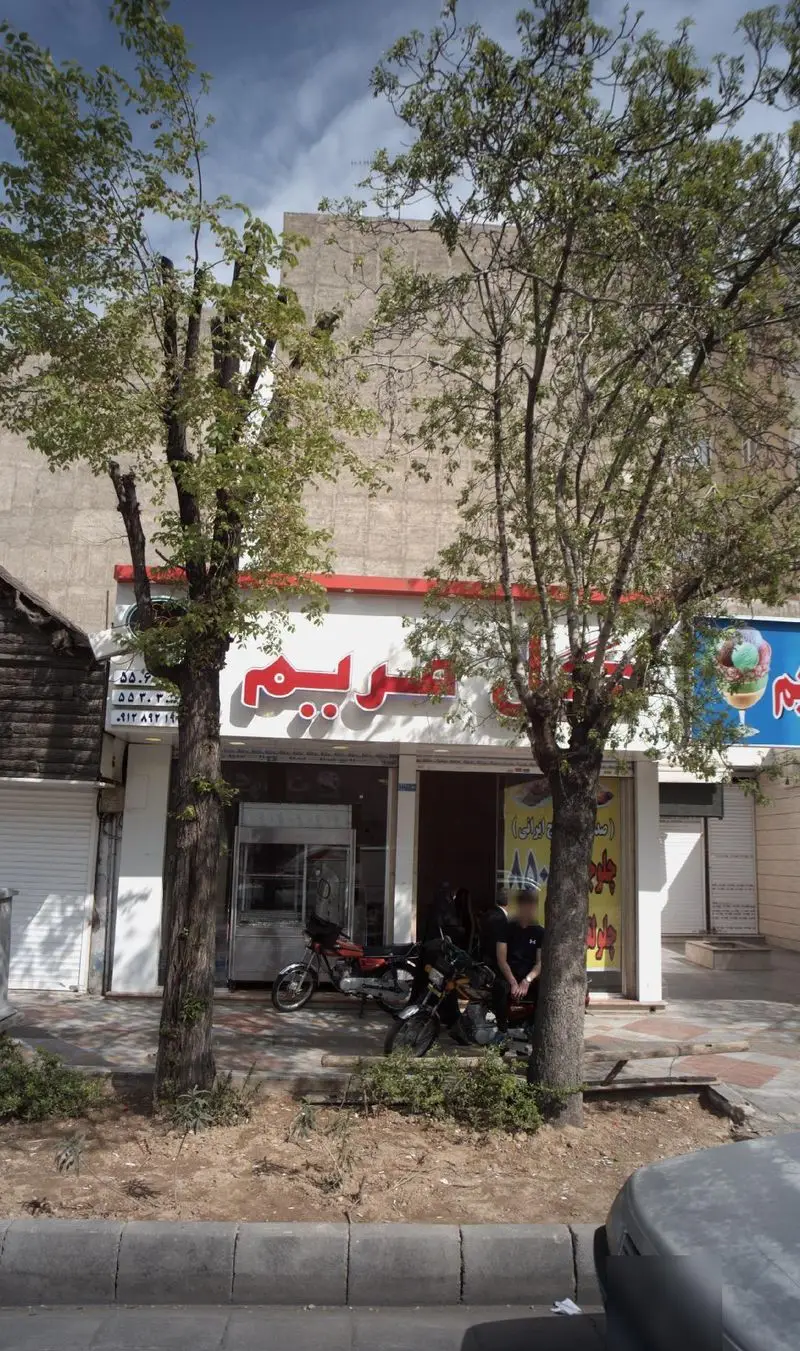 نمای خارجی ویتامینه گل مریم، با تابلوی بزرگ فروش بستنی و ویتامینه، درختان سبز کنار ورودی و موتورسیکلت‌های پارک شده.