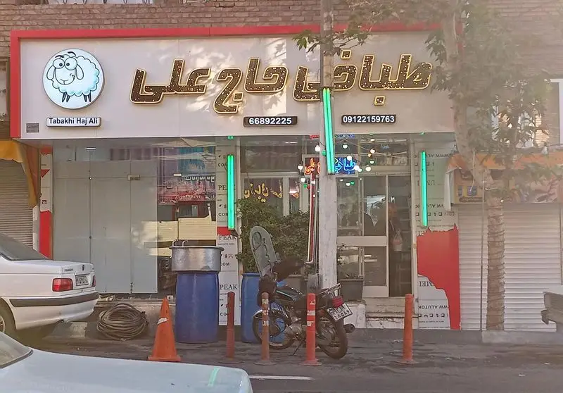 تصویری از سردر طباخی حاج علی، مرکز تهیه خوراک‌های سنتی و کله‌پاچه باکیفیت، شامل لوگوی گوسفند و شماره‌های تماس ۶۶۸۹۲۲۲۵ و ۰۹۱۲۲۱۵۹۷۶۳.