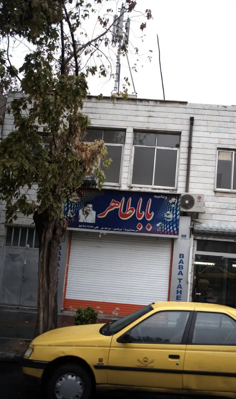 مغازه ویتامینه بابا طاهر، آبمیوه بستنی فروشی در تهران، با تابلوی فارسی جذاب، تصویر مردی و خودروی زرد رنگ در خیابان.
