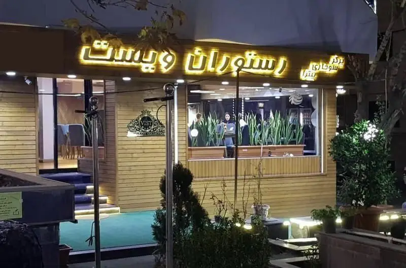 فست فود ویست فود در شب، با تابلوی "رستوران ویست" و پیشنهاد برگر، پیتزا. نمای دنج و چوبی با نورپردازی گرم و محیط داخلی سبز.