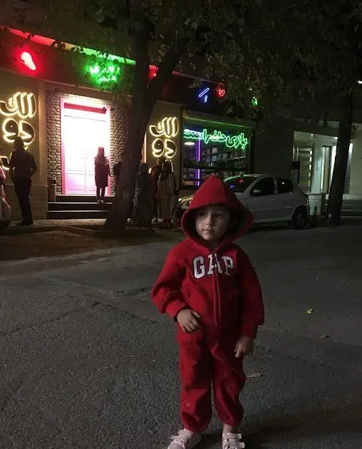 نمایی شبانه از کافه رستوران الک دولک با تابلوهای نئونی رنگارنگ. کودکی با لباس قرمز و لوگوی گپ در پیش‌زمینه، فضایی شاد و مناسب بازی و سرگرمی.