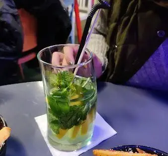 نوشیدنی موهیتو معطر، تازه و خنک با برگ‌های سبز نعناع و تکه‌های لیمو در لیوان بلند روی میز کافه رستوران الک دولک، محیطی آرامش‌بخش و دلپذیر.