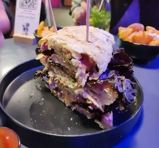 ساندویچ غول‌پیکر و لایه لایه "کافه رستوران الک دولک" با نان خوش‌طعم و مواد پرملات، کنار سیب‌زمینی سرخ کرده، تجربه غذایی لذت‌بخش در محیط دنج کافه.