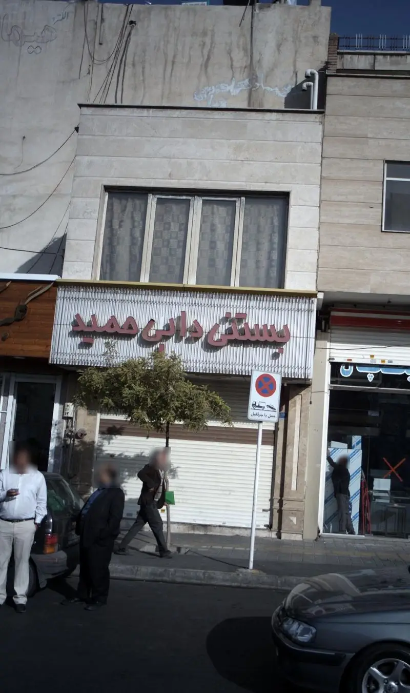 نمای بیرونی بستنی دایی حمید در یک روز آفتابی با تابلوی قرمز، نشان‌دهنده کسب‌وکار فعال و پر جنب‌وجوش در خیابان.