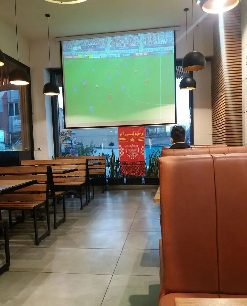 فضای داخلی رستوران ویانا با میز و صندلی‌های راحت، نمایش بازی فوتبال و بنر پرسپولیس، مکانی عالی برای دورهمی و لذت بردن.