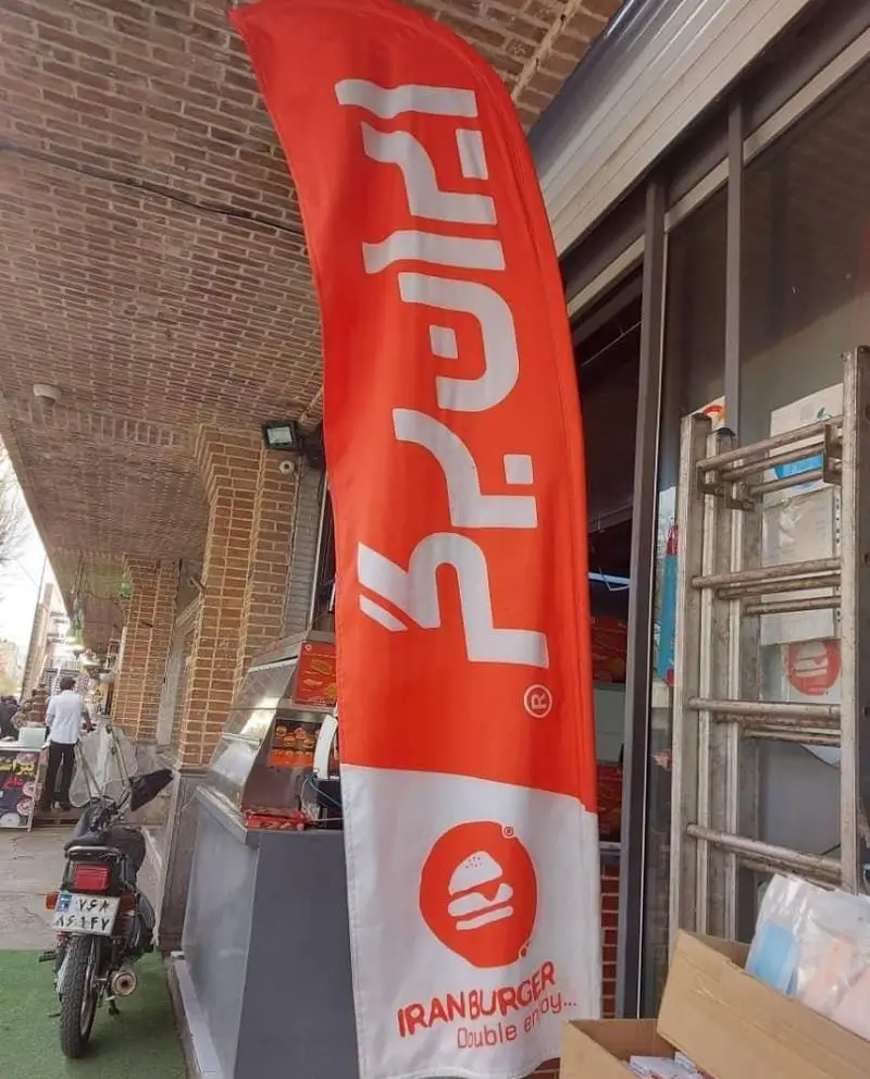 تصویر پرچم قرمز و سفید ایران برگر (سرزمین سوخاری) در جلوی یک شعبه فست فود با موتور پارک شده در پیاده رو.