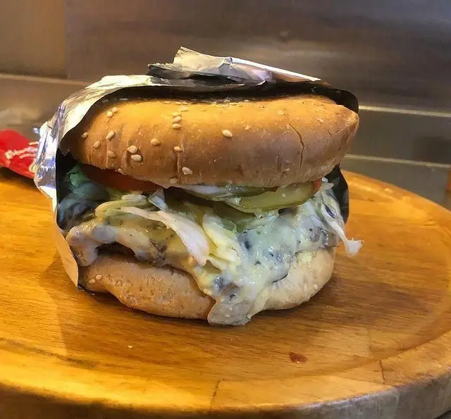 تصویری جذاب از همبرگر ویژه فست فود هیزم با نان کنجدی، پنیر ذوب شده، کاهو و سس مخصوص، آماده سرو روی سینی چوبی.