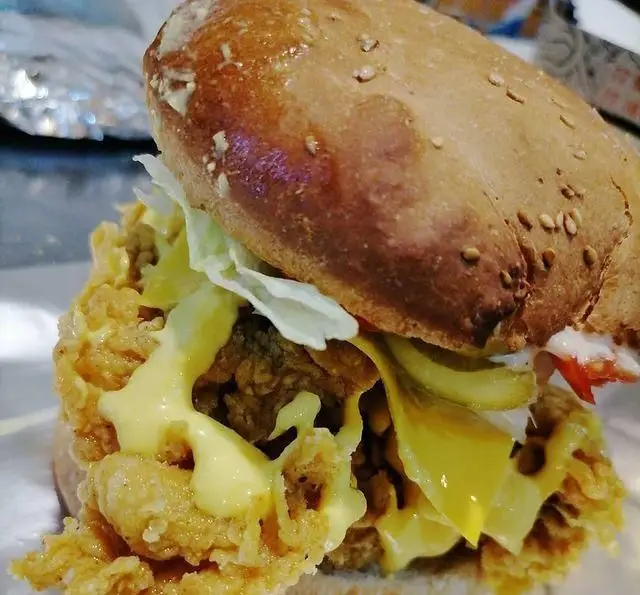 یک همبرگر بزرگ و اشتها آور از فست فود هیزم با فیله مرغ سوخاری، پنیر ذوب شده، کاهو، خیارشور و سس سفید روی نان کنجدی.