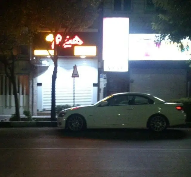 تصویر شبانه از نمای بیرونی فست فود هیزم با تابلوی نئون قرمز روشن، در کنار خیابان و یک خودروی سفید رنگ پارک شده روبرو