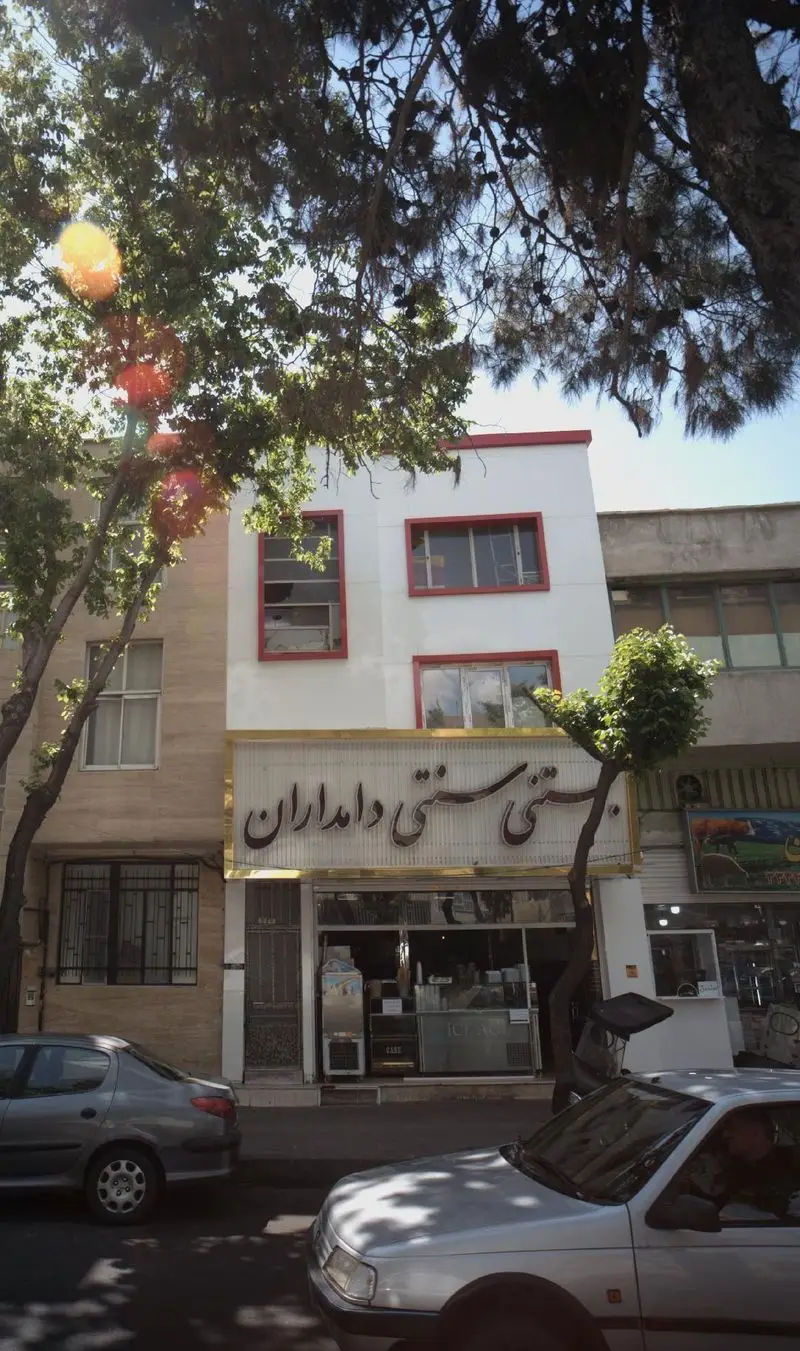 نمای بیرونی بستنی سنتی دامداران، مغازه بستنی‌فروشی با تابلوی روشن، درختان سبز و خودروها در خیابان، یک روز آفتابی.